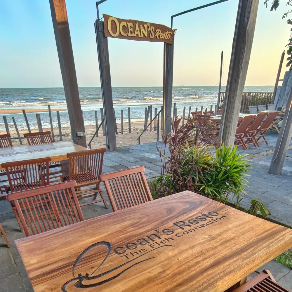 Meja kayu berukir logo Ocean's Resto The Fish Connection dengan laut di latar saat senja